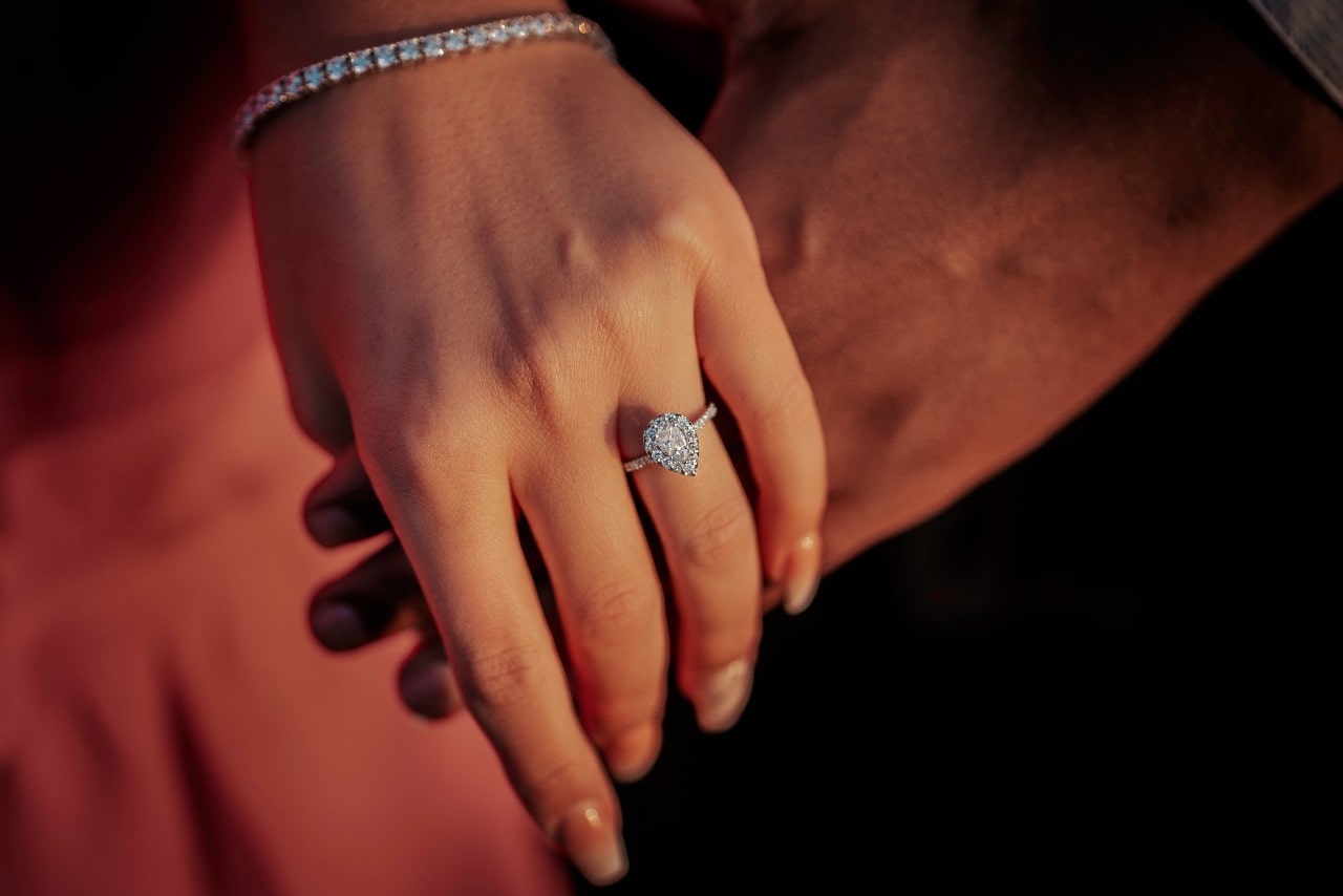 a couple holding hands, the woman wearing a white gold, halo pear shape engagement ring