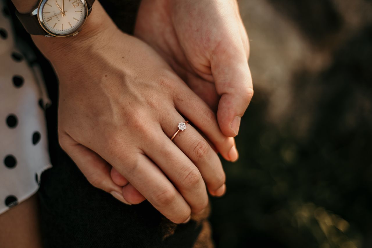 A woman and her fiance subtly hold hands to show of her gold solitaire engagement ring.