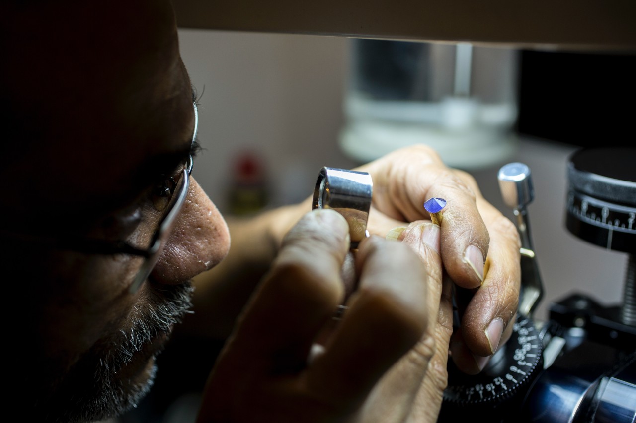 A close-up of a jeweler with a magnifying glass looking at a blue gemstone. 