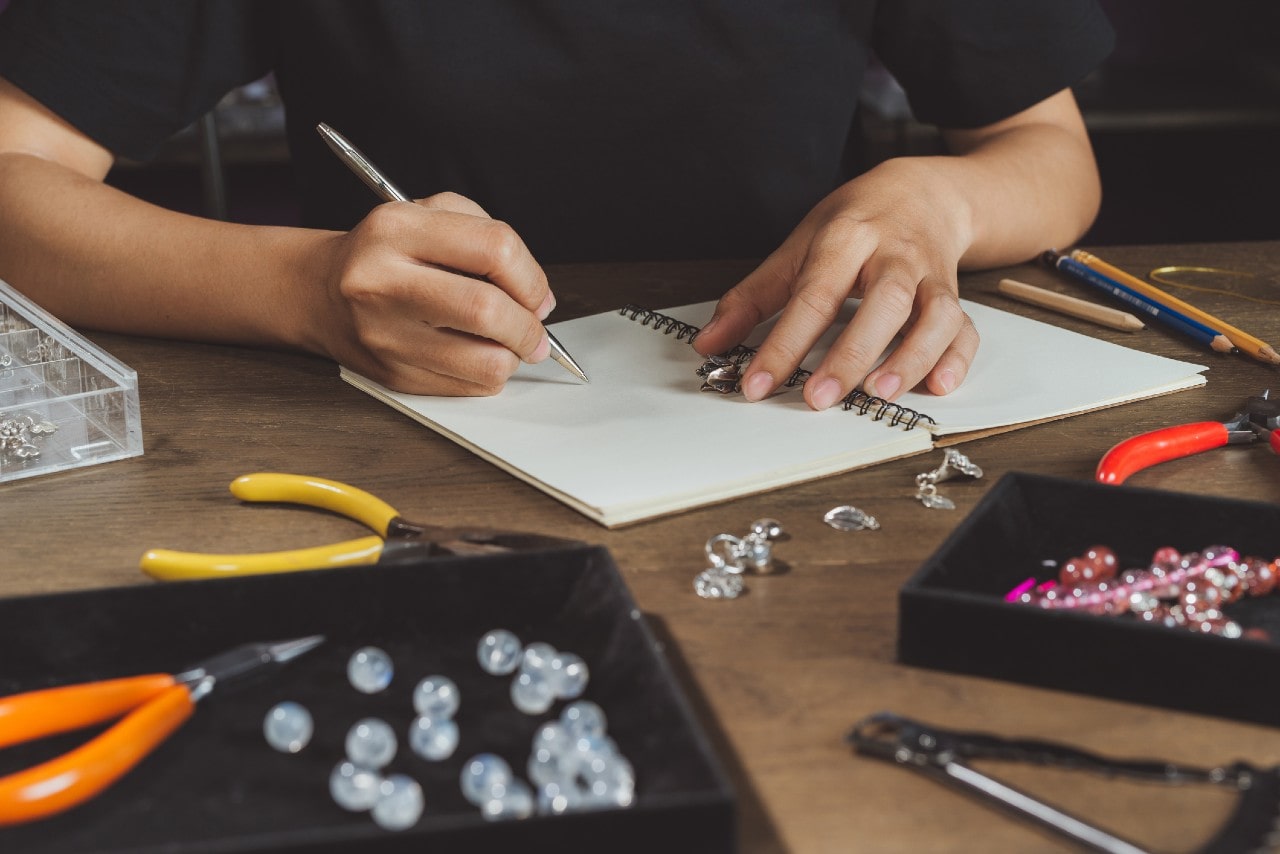 A jewelry designer sketches a design by hand in a notebook surrounded by tools, beads, and metal components on a wooden workbench.