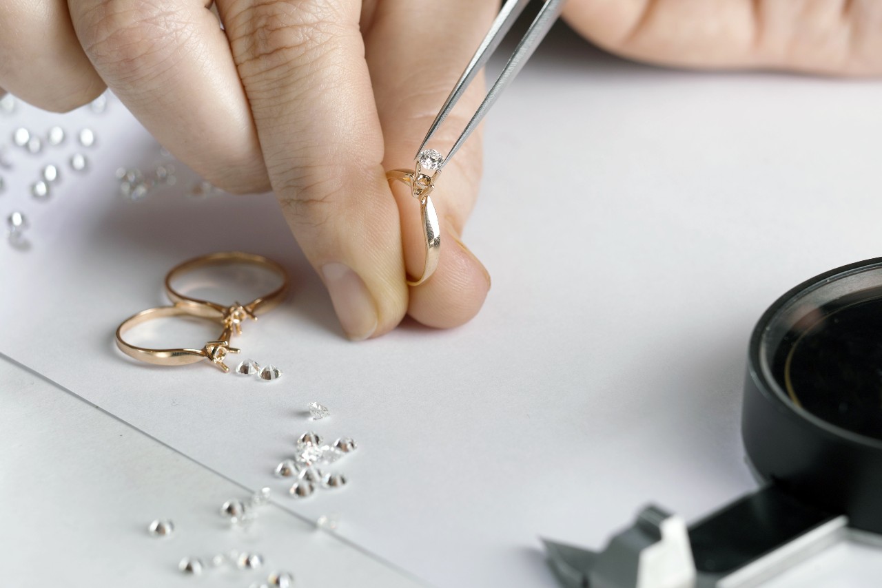 A close-up of a jeweler with tweezers placing a diamond onto the yellow gold solitaire engagement ring setting.