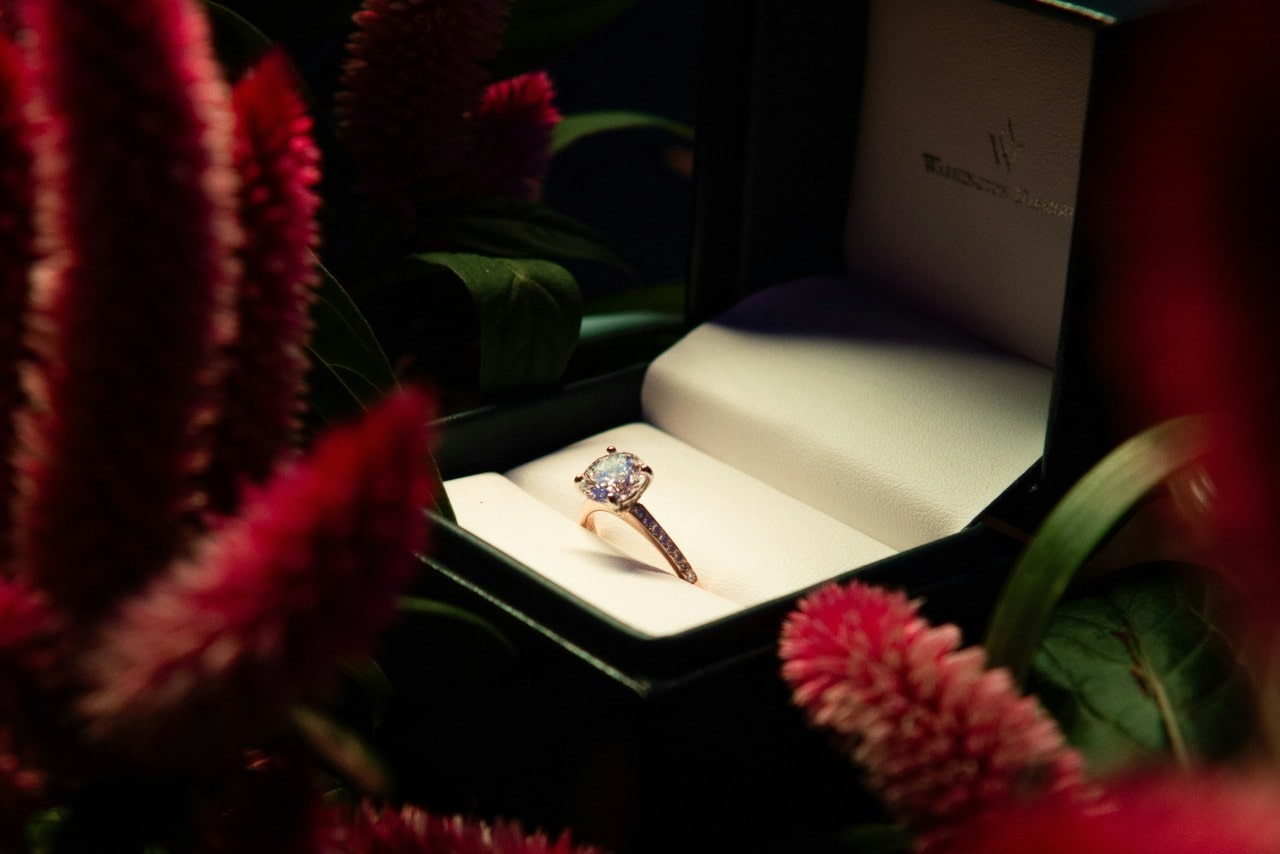 A close up of a round-cut diamond engagement ring in a rose gold setting with a pavé band, displayed in a green jewelry box surrounded by pink flowers.
