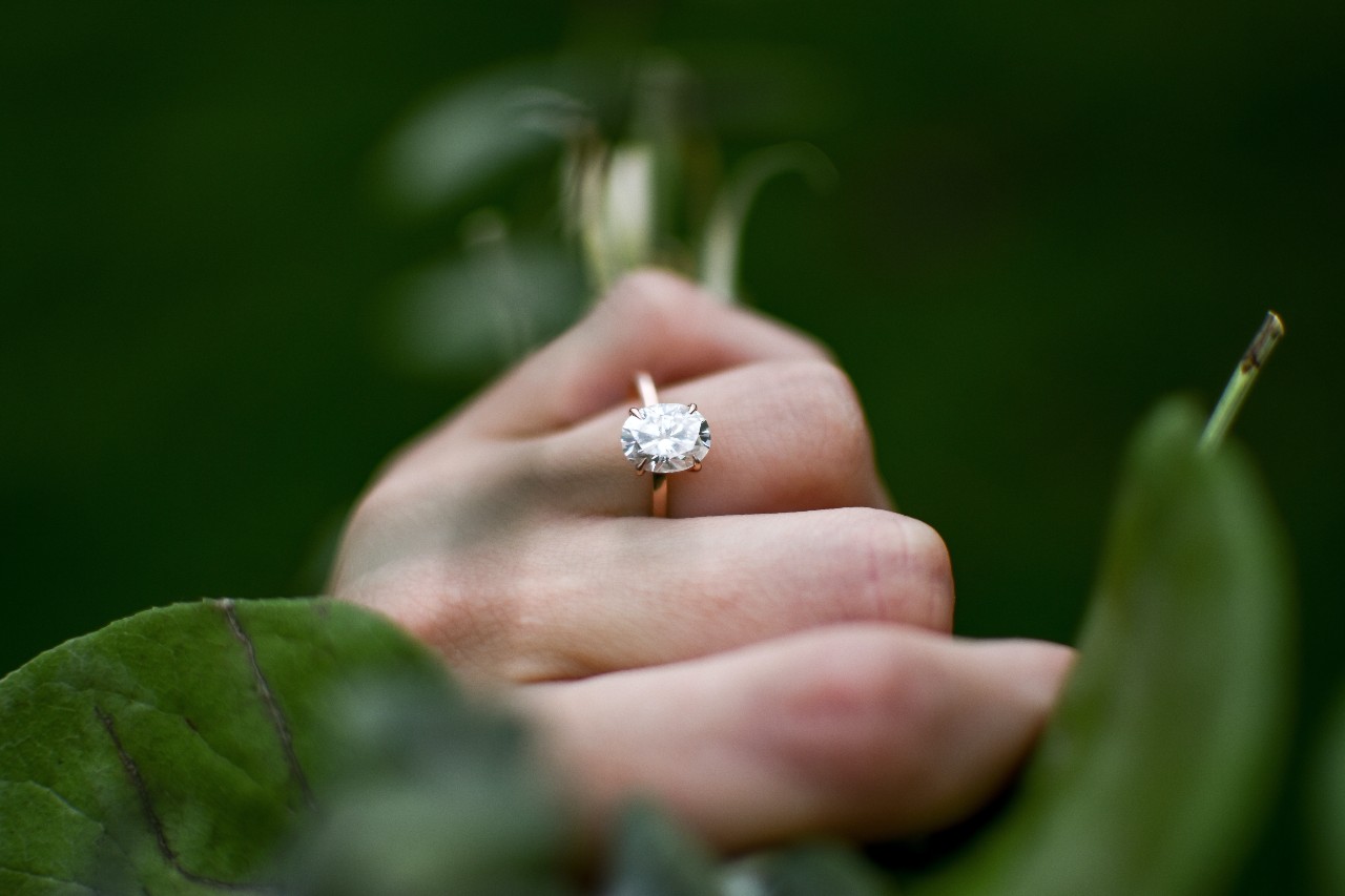 close up image of a hand wearing a gold band engagement ring surrounded by greenery