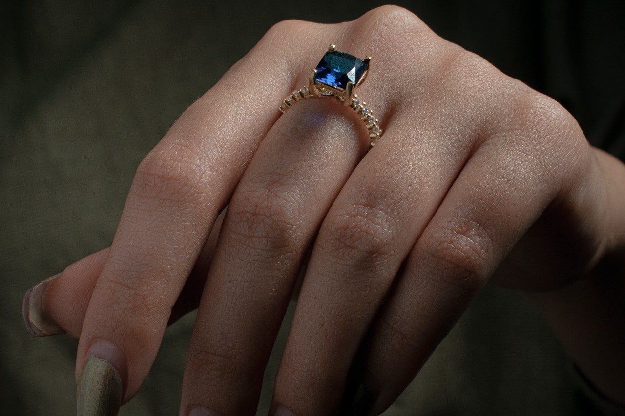 close up image of a woman’s hand wearing a yellow gold fashion ring with a princess cut sapphire