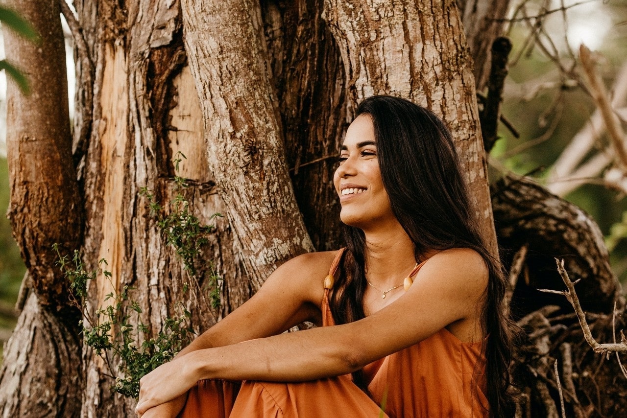 smiling lady sitting up against a tree and wearing a fashion necklace