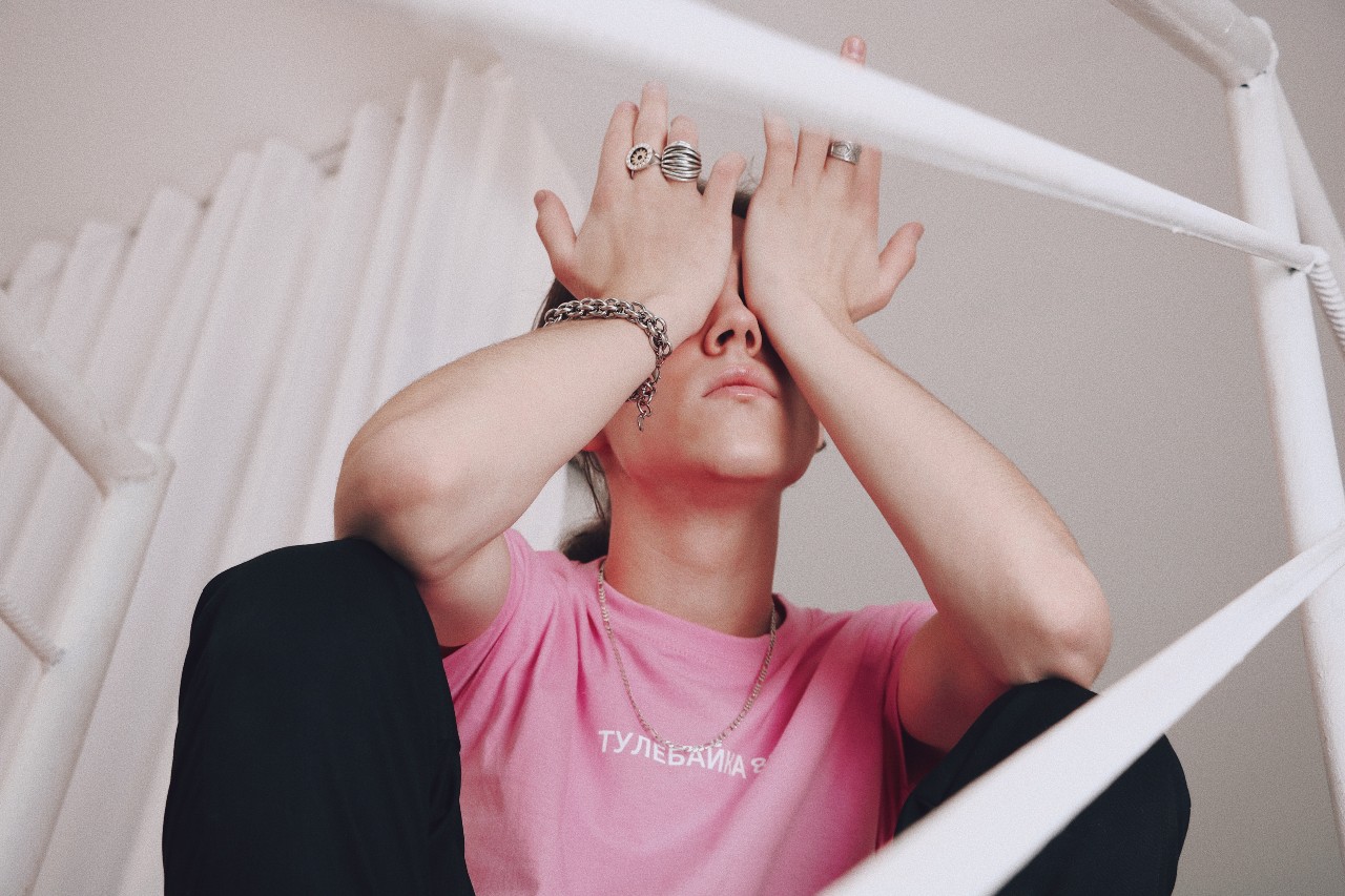 A woman in a pink T-shirt covers her face with both hands adorned with chunky silver stacking rings and a chain bracelet, sitting on a white staircase.