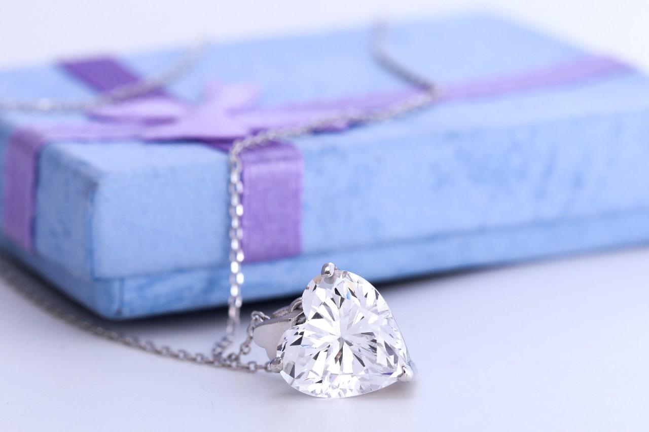 A heart-shaped diamond necklace draped over a closed, light blue gift box with a purple ribbon.
