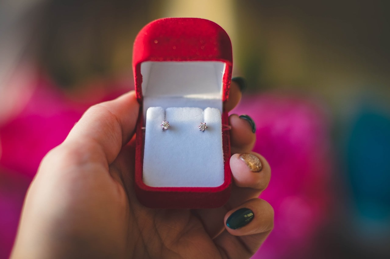 A close-up of a hand holding an open red velvet jewelry box containing sparkly diamond stud earrings, set against a blurred, colorful background.