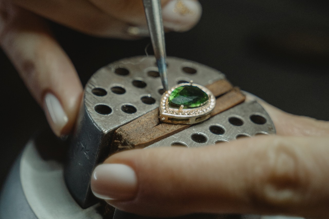 A jeweler's hands carefully set a vibrant green gemstone into a diamond-encrusted setting using a precision tool under a microscope
