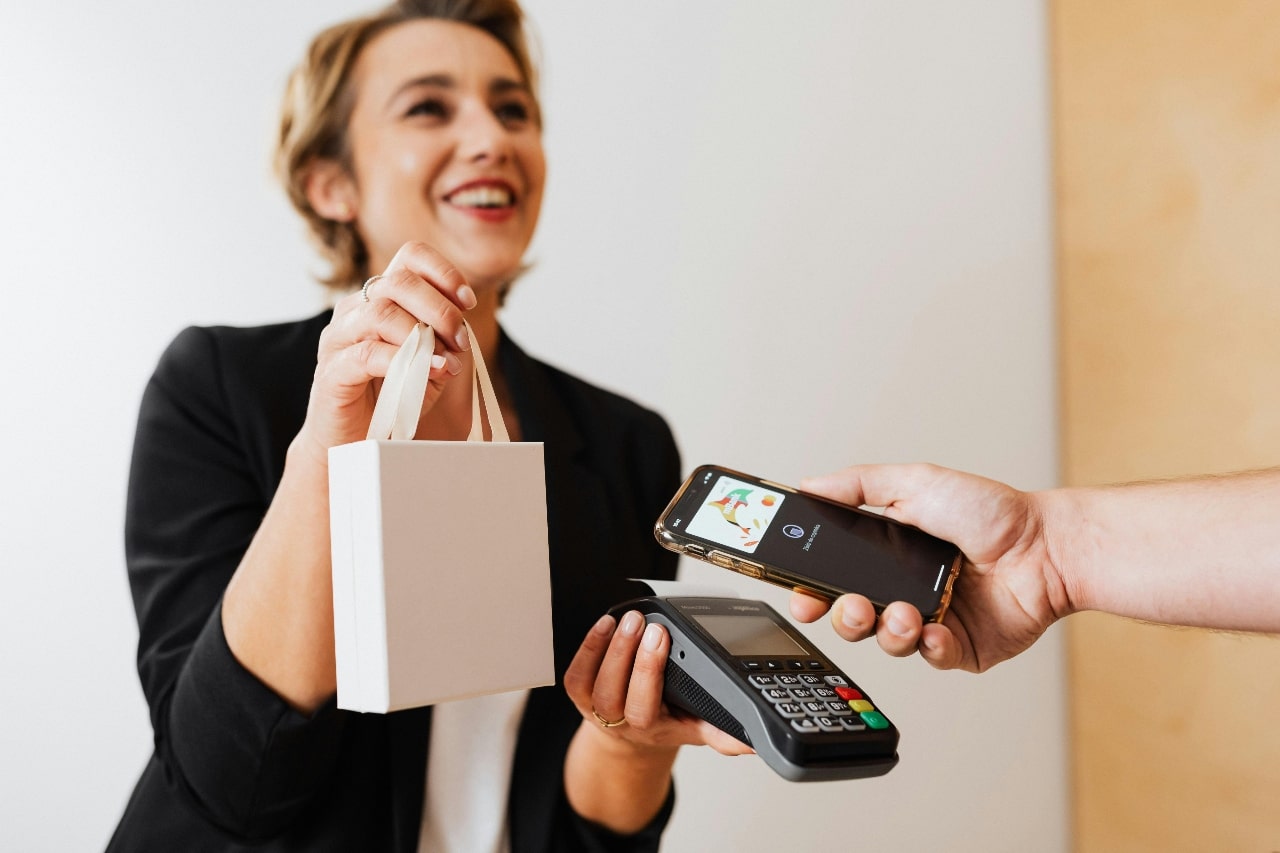 A smiling saleswoman hands a small shopping bag to a customer who is making a contactless payment with a smartphone.