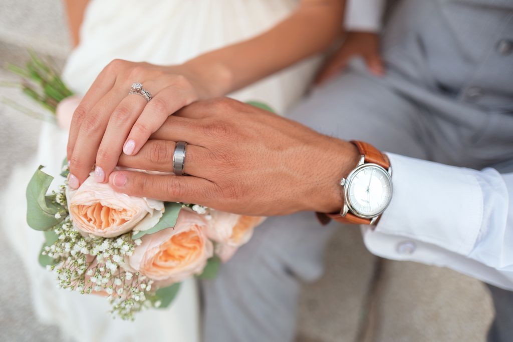 A bride and groom with their hands on top of the bride’s pale peach rose bouquet; focus on the gunmetal grey wedding band with a matte finish on the groom’s ring.