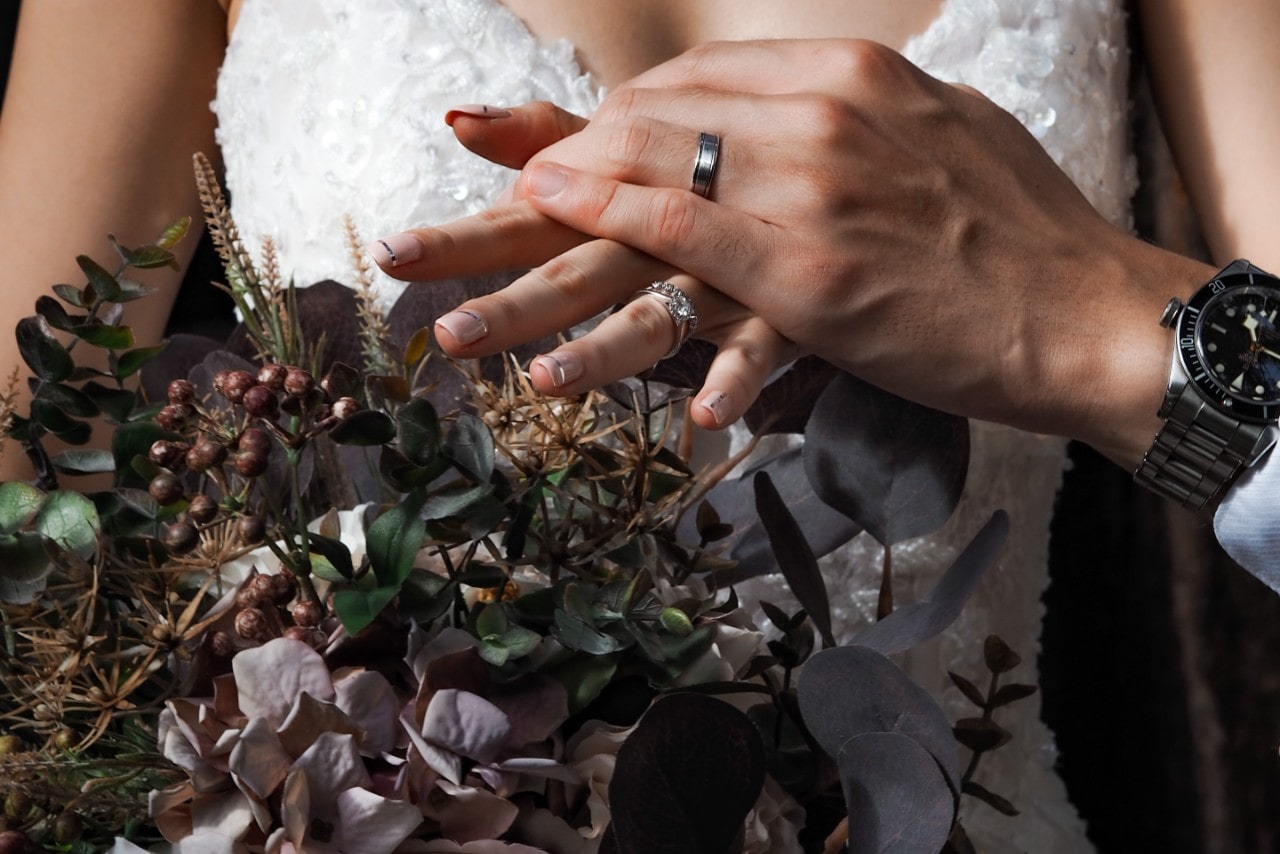 a bride and groom holding hands over her winter boutique showing her diamond wedding band and engagement ring along with his wedding band and watch
