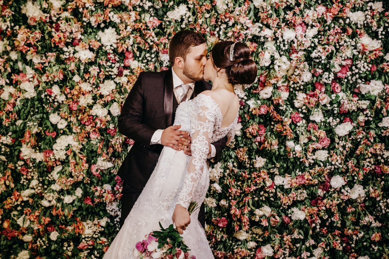 A bride and groom kissing in front of a wall of pink and white flowers with greenery.