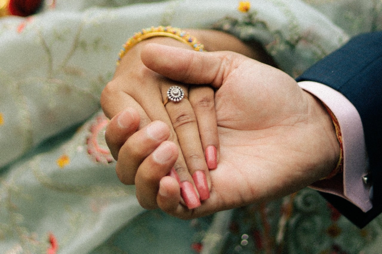 A close-up of two hands gently holding, one wearing a floral ring, with the background featuring soft, embroidered fabric. A close-up of two hands gently holding, one wearing a floral ring, with the background featuring soft, embroidered fabric.
