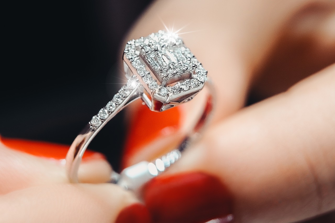 Close-up of a diamond ring with a square halo setting held between fingers with red nails. Close-up of a diamond ring with a square halo setting held between fingers with red nails.