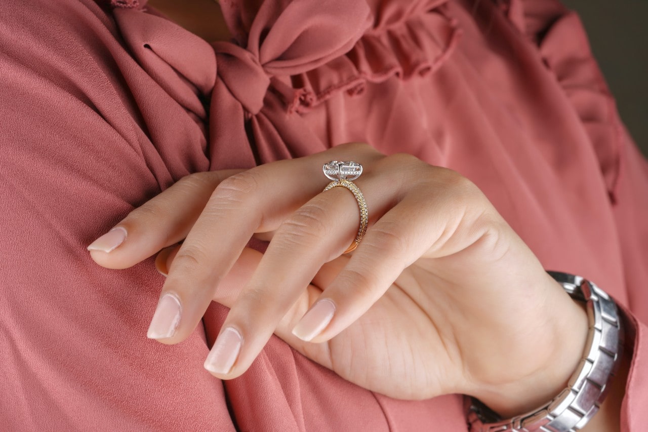 a woman in a pink blouse raising her hand to her chest and wearing an emerald cut engagement ring