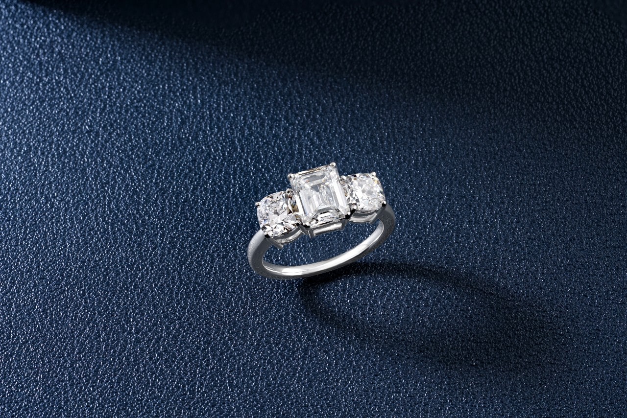 a three stone, emerald cut engagement ring against a black textured background