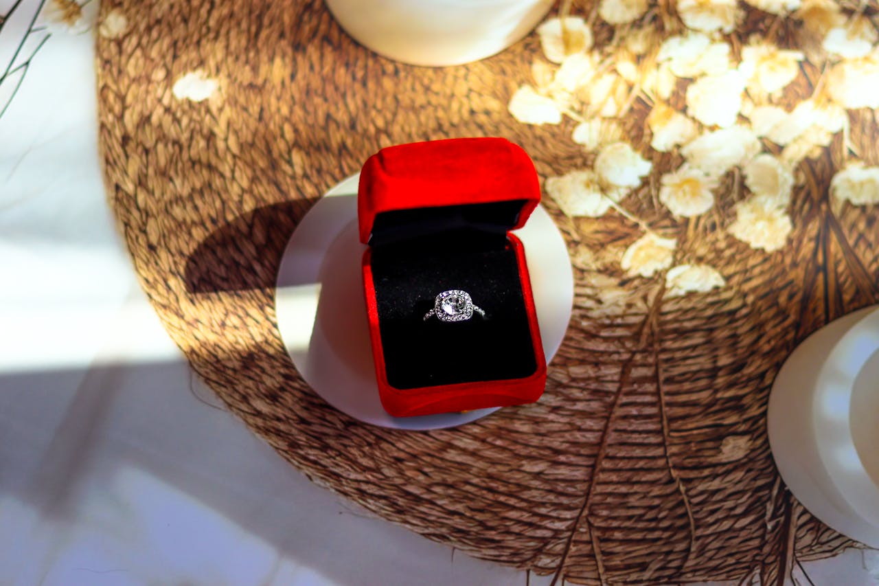 A close-up of a white gold halo engagement ring inside a black and red velvet jewelry box on a round woven mat.