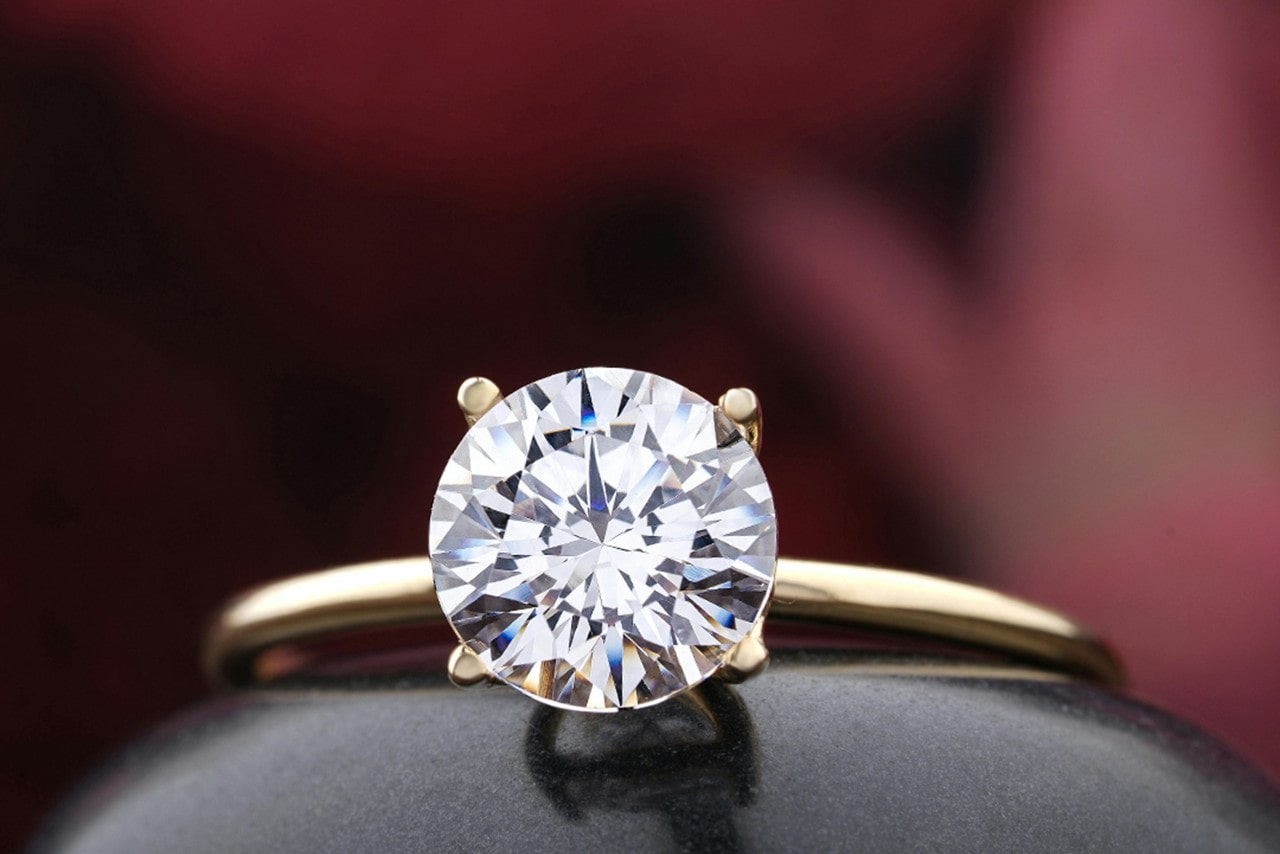 A yellow gold solitaire engagement ring with round cut diamond against a burgundy blurred background.