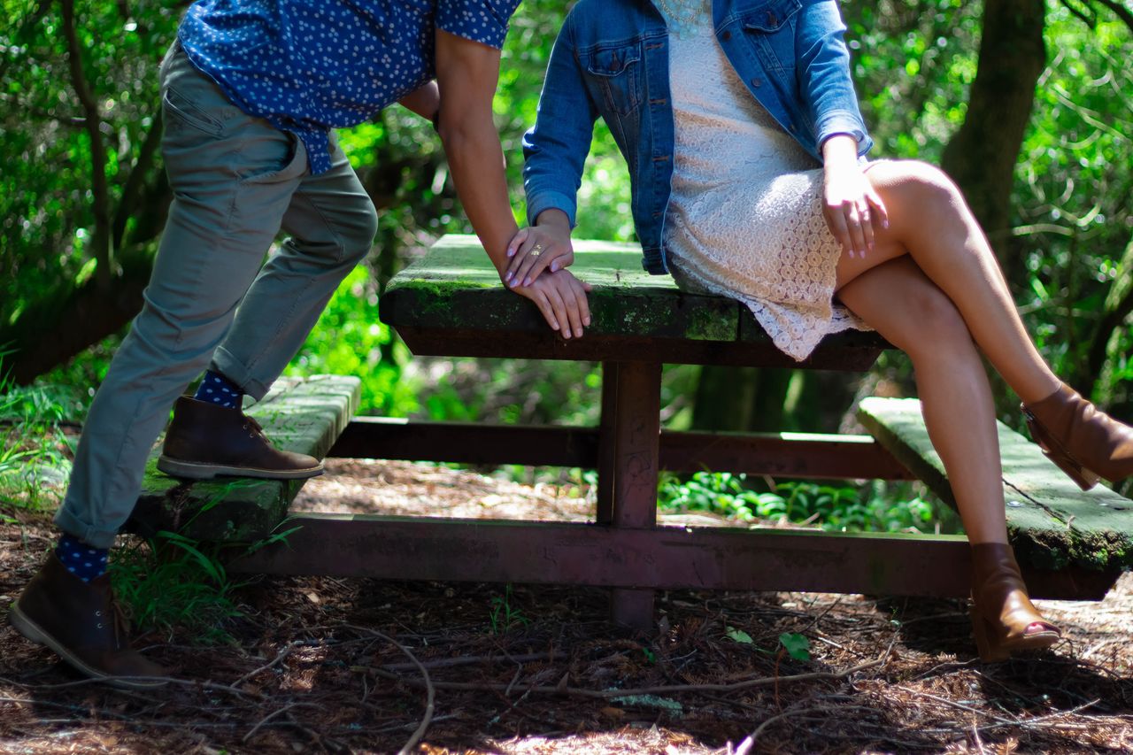 An engaged couple kisses on a moss-covered picnic table in a state park.