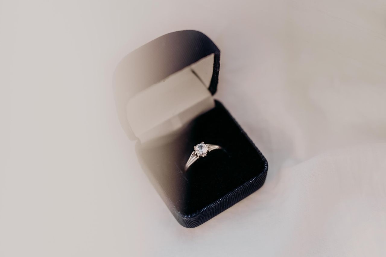 A solitaire engagement ring sits in a black box on white, sheer fabric.