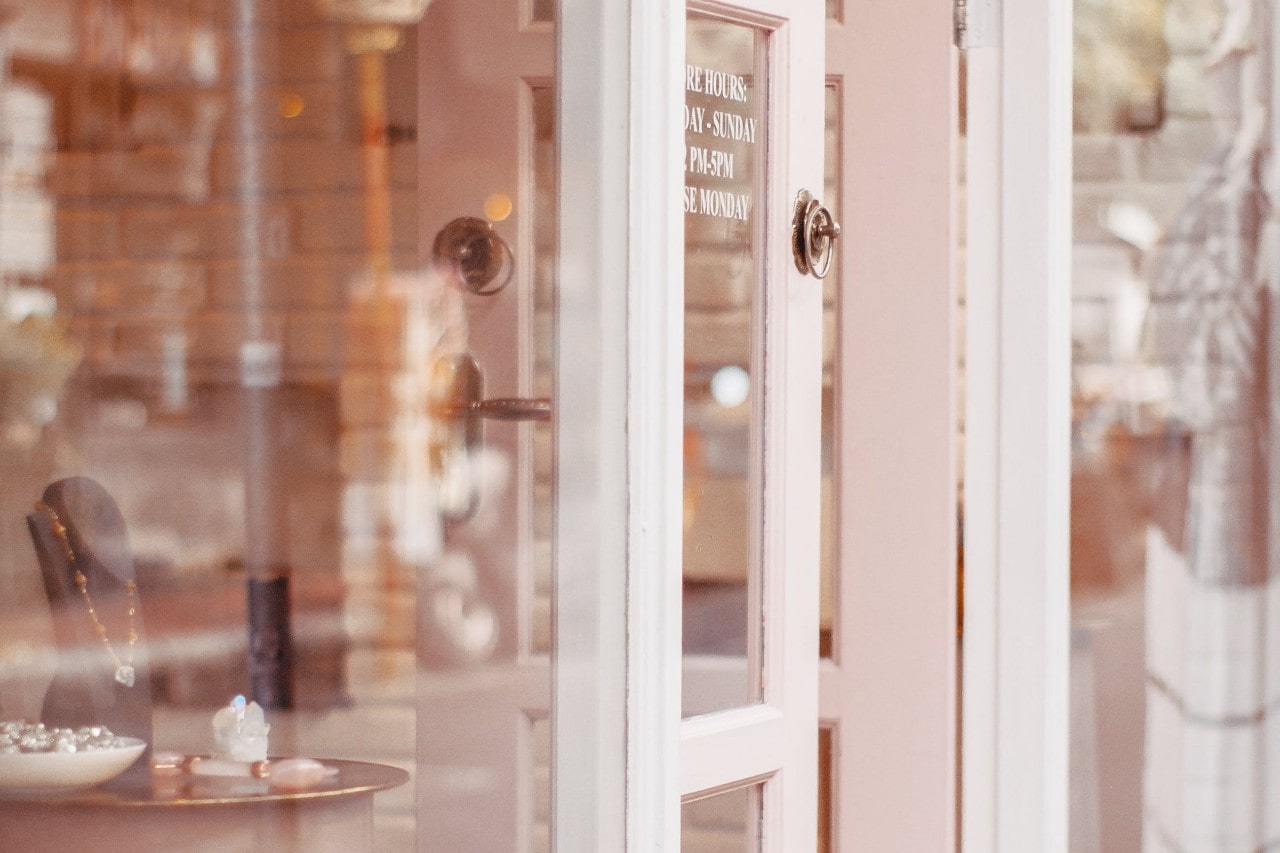 Open shop door with pink frame and glass, reflecting jewelry display of necklaces and rings inside.