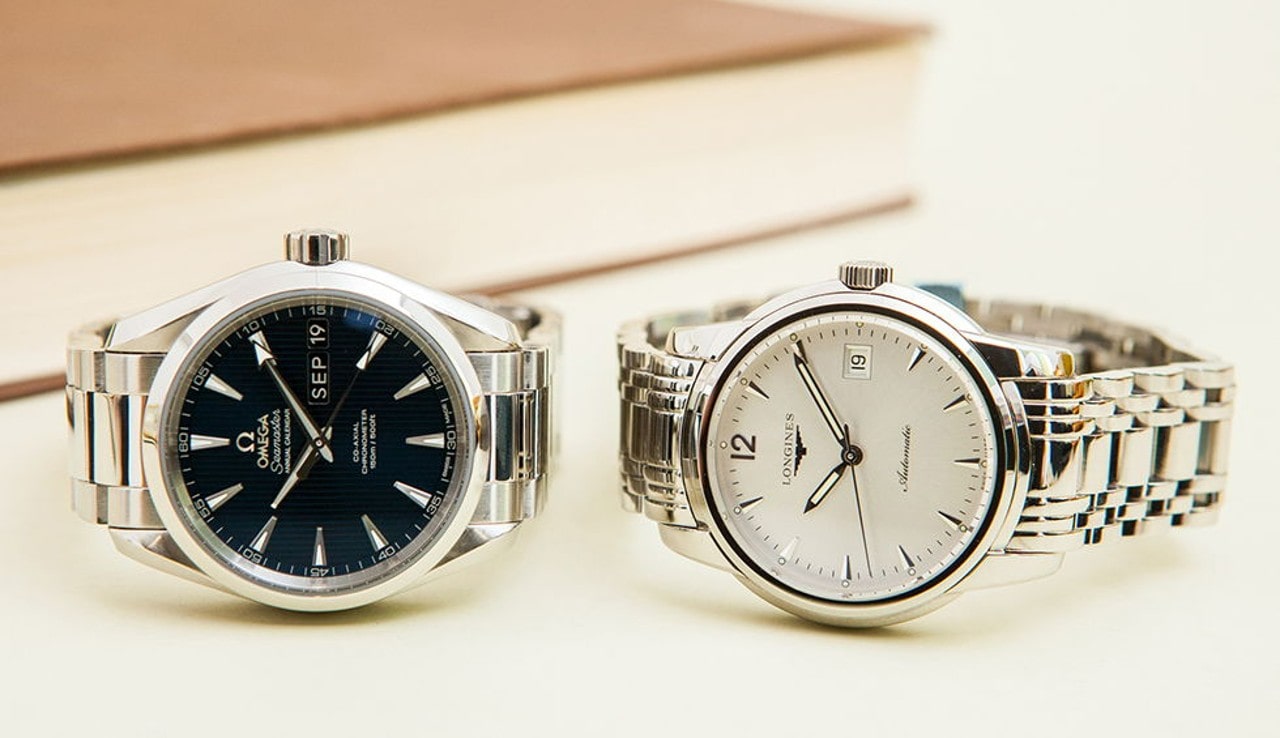 A side-by-side display of an Omega Seamaster with a black dial and a Longines automatic watch with a white dial, both featuring stainless steel bracelets. A side-by-side display of an Omega Seamaster with a black dial and a Longines automatic watch with a white dial, both featuring stainless steel bracelets.