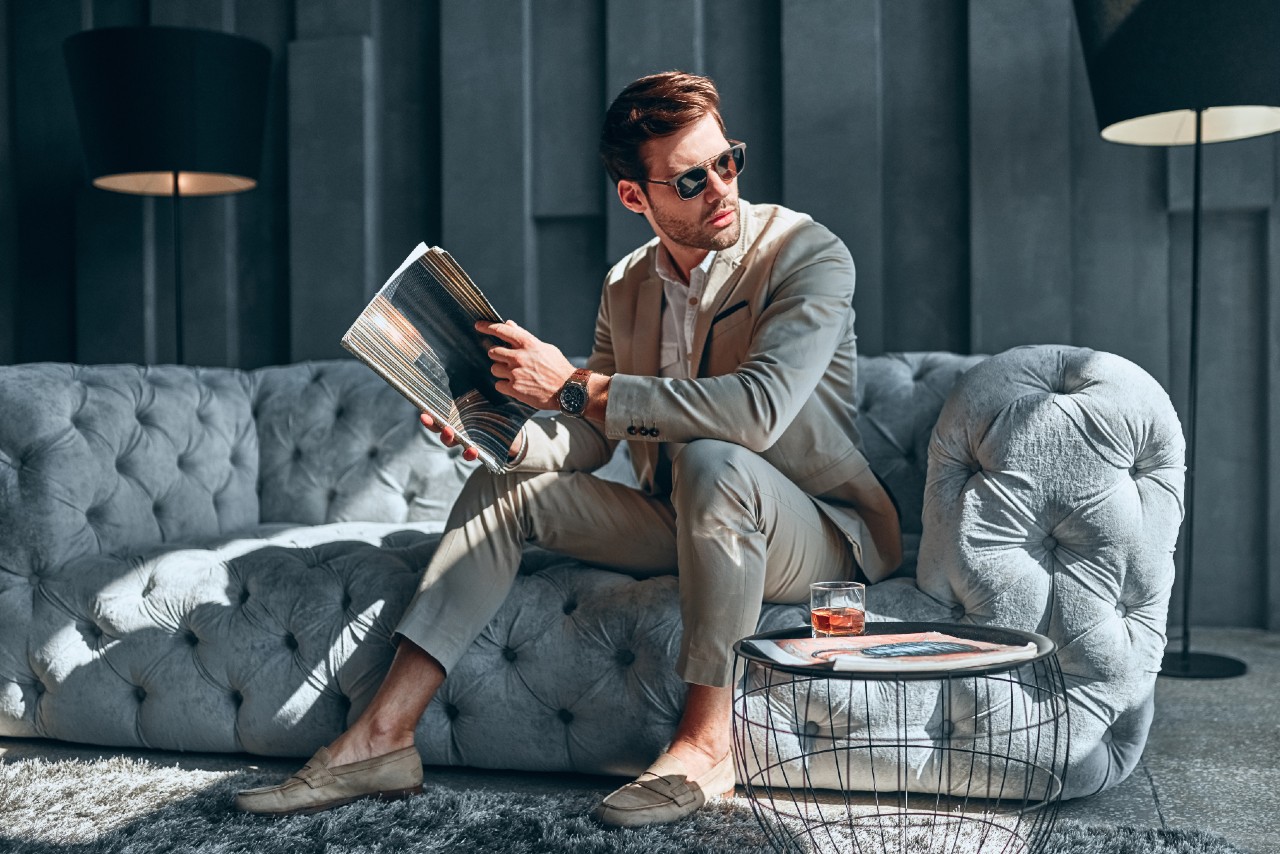 A man in a beige suit and sunglasses sits on a tufted sofa while reading a magazine, showcasing a luxury leather watch on his wrist. A man in a beige suit and sunglasses sits on a tufted sofa while reading a magazine, showcasing a luxury leather watch on his wrist.