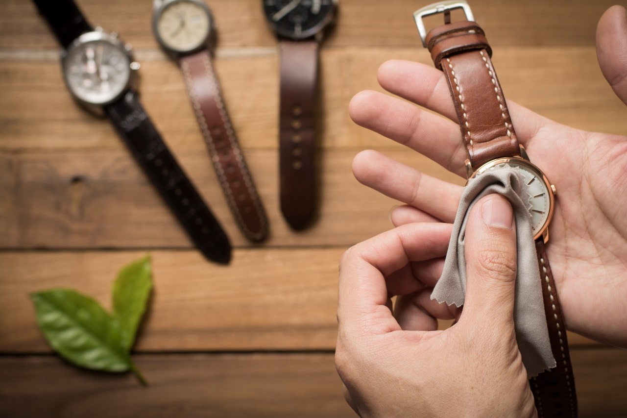 A close up of a man wiping a leather wristwatch with a microfiber piece of cloth while three other watches rest on a wooden surface in the background A close up of a man wiping a leather wristwatch with a microfiber piece of cloth while three other watches rest on a wooden surface in the background