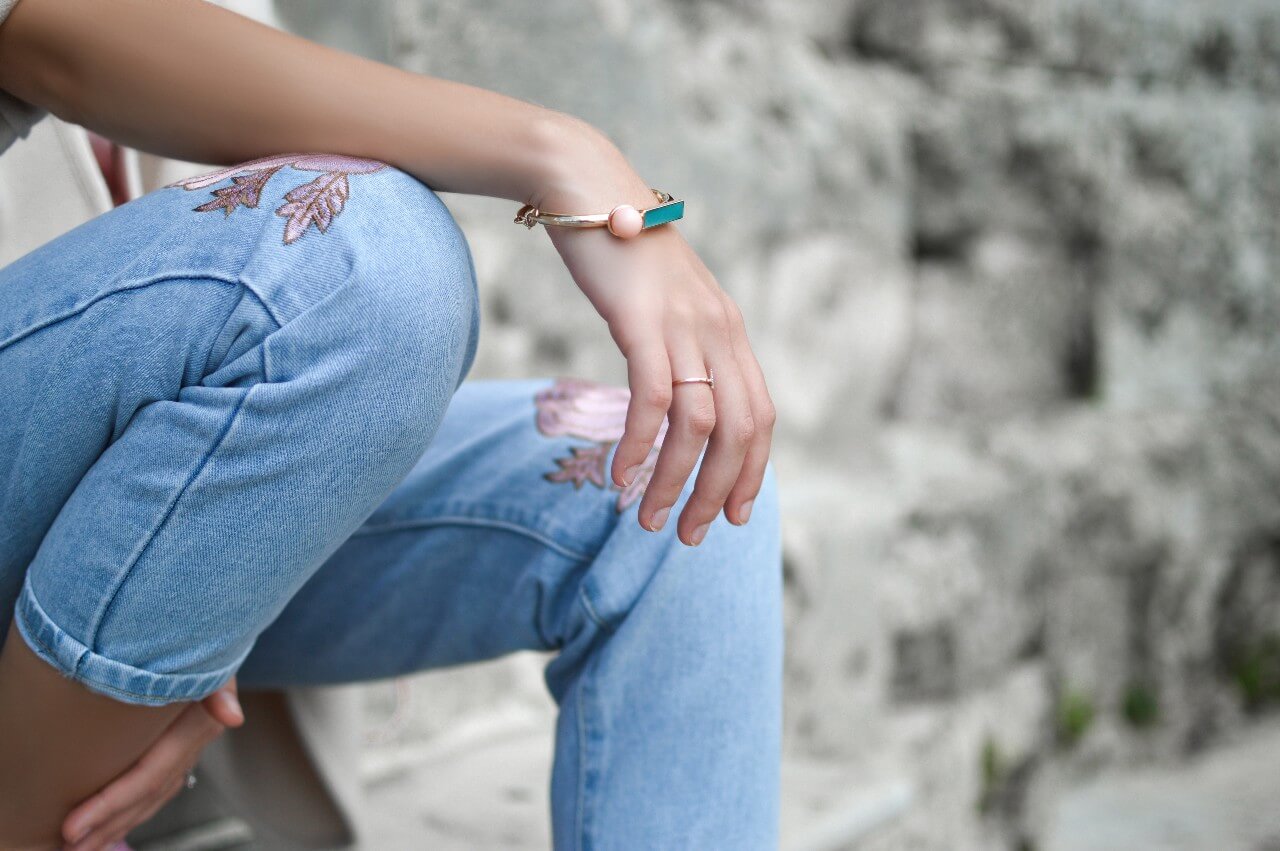 A woman in embroidered jeans rests her arm on her knee to show off her gemstone bracelet.