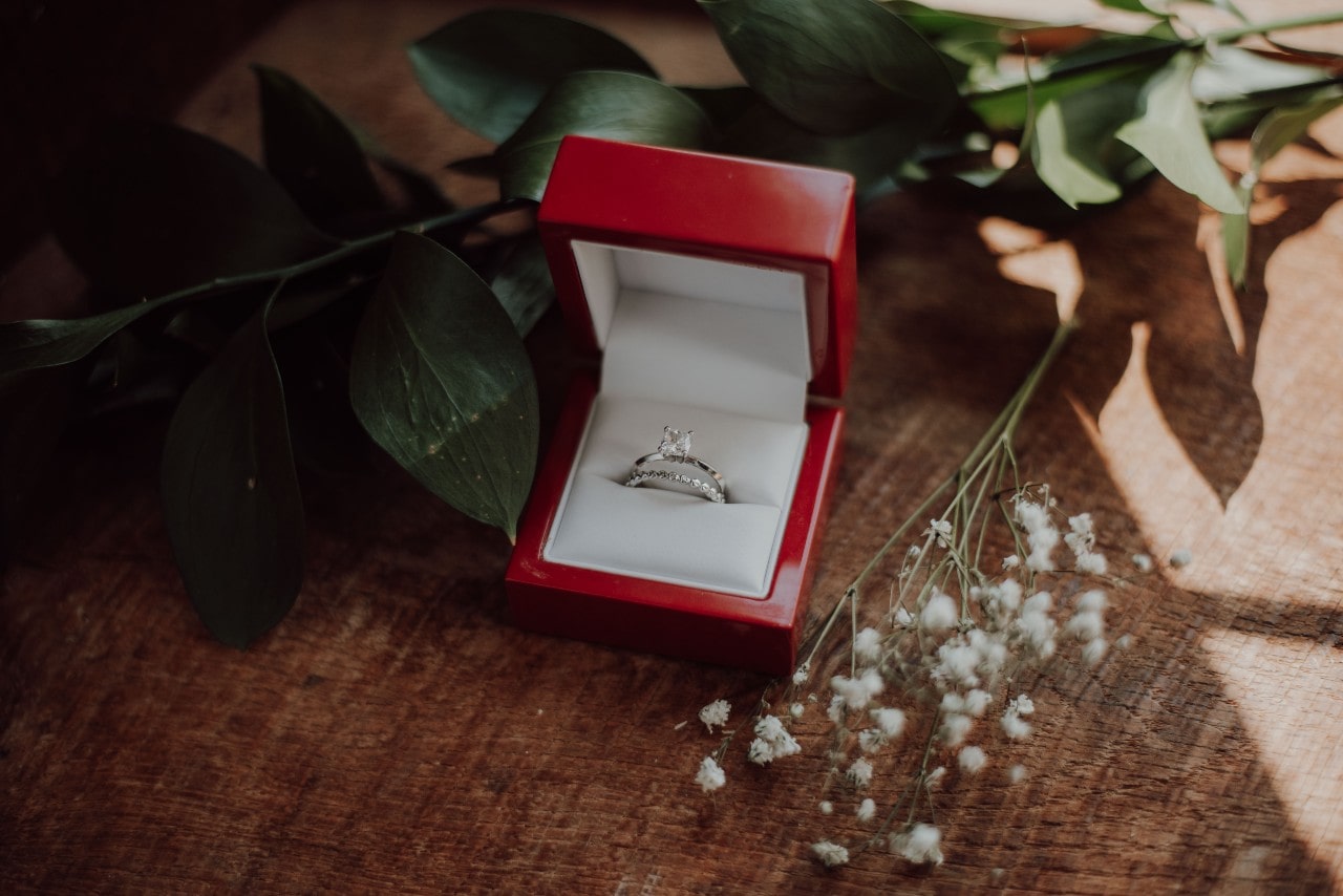 A red jewelry box with a white interior holds a sparkling diamond engagement ring and a matching wedding band, placed on a wooden surface. A red jewelry box with a white interior holds a sparkling diamond engagement ring and a matching wedding band, placed on a wooden surface.