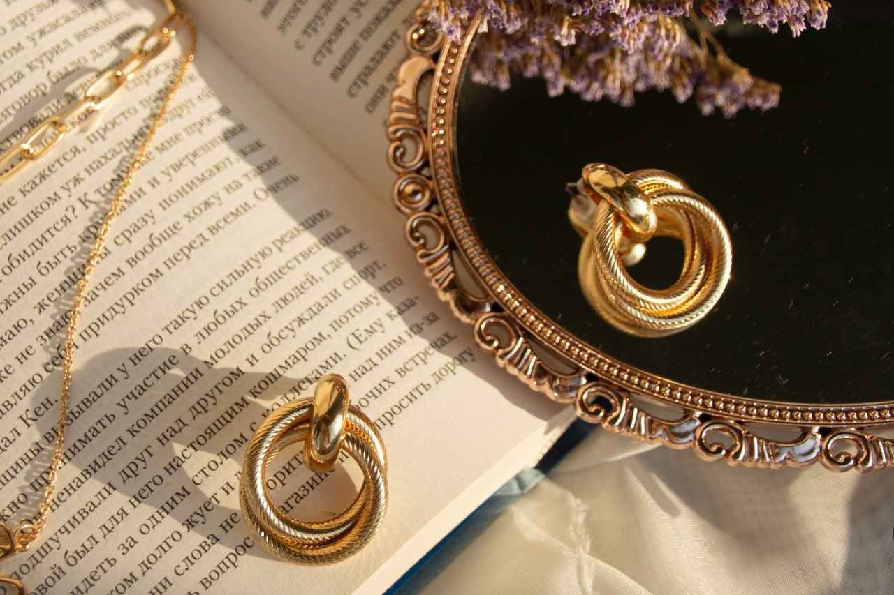 A yellow gold necklace and a twisted ring placed on an open book, with a vintage mirror reflecting the ring and dried lavender flowers A yellow gold necklace and a twisted ring placed on an open book, with a vintage mirror reflecting the ring and dried lavender flowers
