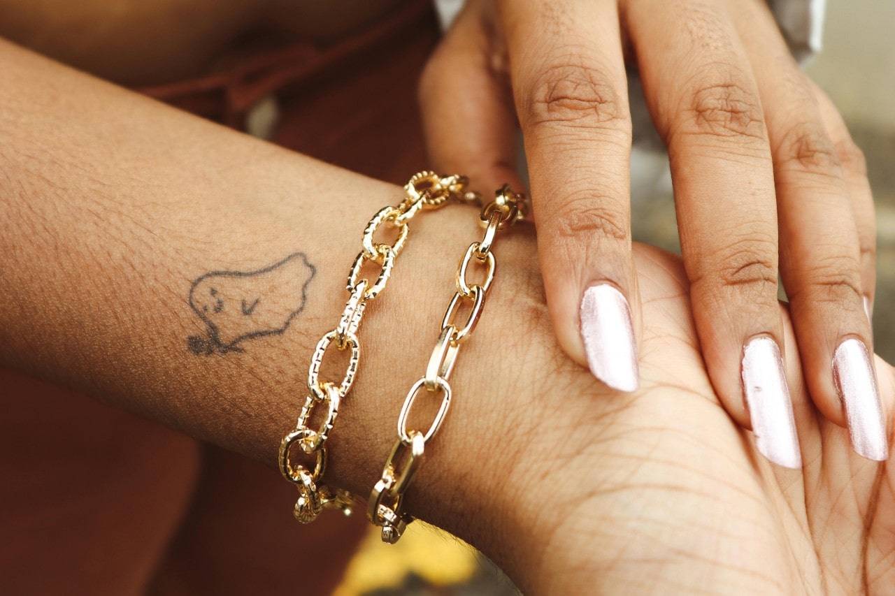 A woman’s manicured hand touching her wrist, adorned with two gold chain bracelets below a small ghost tattoo.