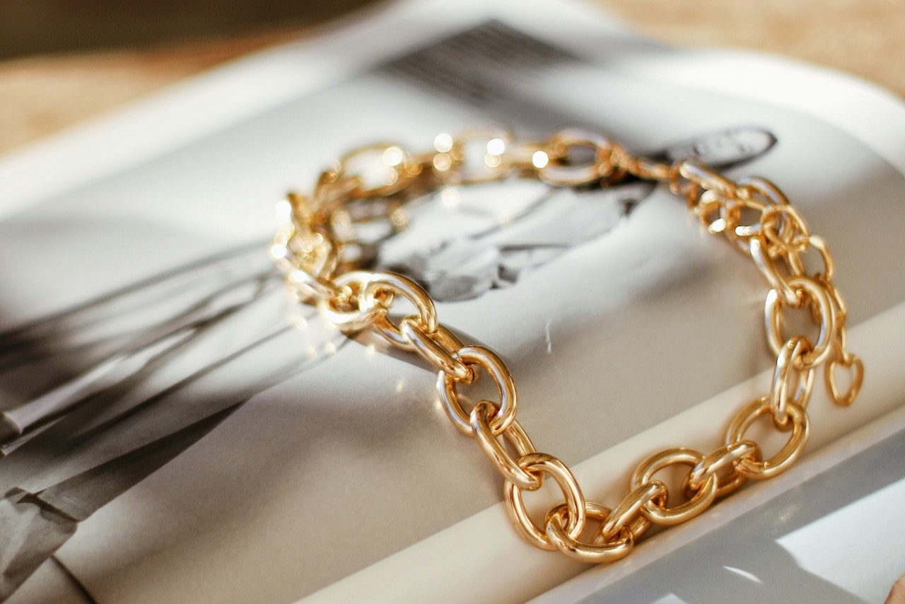 A gold chain bracelet with large, interlocking links resting on an open magazine with a black-and-white fashion photograph.