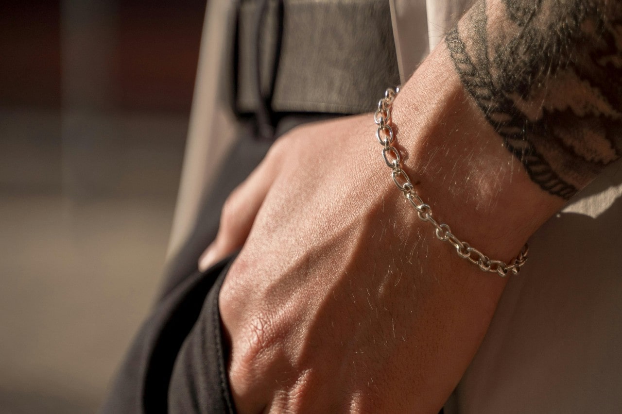 A close-up of a person's wrist wearing a silver chain bracelet, with a tattoo partially visible and their hand resting in a pocket.
